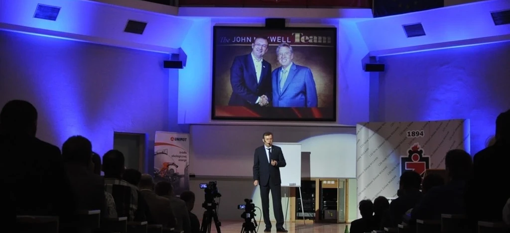 A spokesperson in a suit giving a speech at an event while standing at a podium with a display device and electronic devices in the background. Clothing includes a coat and outerwear. QUNIMOT 2016 presentation.