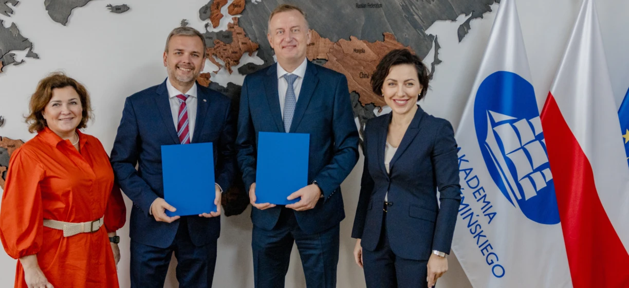 Pracownik umysłowy w garniturze i marynarce uśmiechający się przed flagami Grenlandii, Federacji Rosyjskiej, Mongolii i Chin. Loga uniwersytetów w tle.