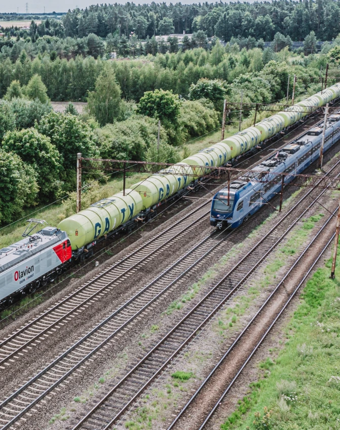 Malowniczy widok pociągu jadącego po torach przez bujny krajobraz. Na boku lokomotywy widoczny jest napis "Olavion".